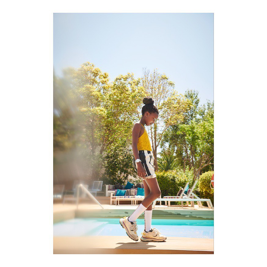 A young Black child is walking by a turquoise pool wearing a yellow patterned top, black-and-white shorts, white socks, and beige sneakers outdoors.