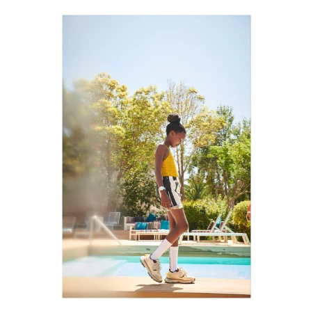 A young Black child is walking by a turquoise pool wearing a yellow patterned top, black-and-white shorts, white socks, and beige sneakers outdoors.