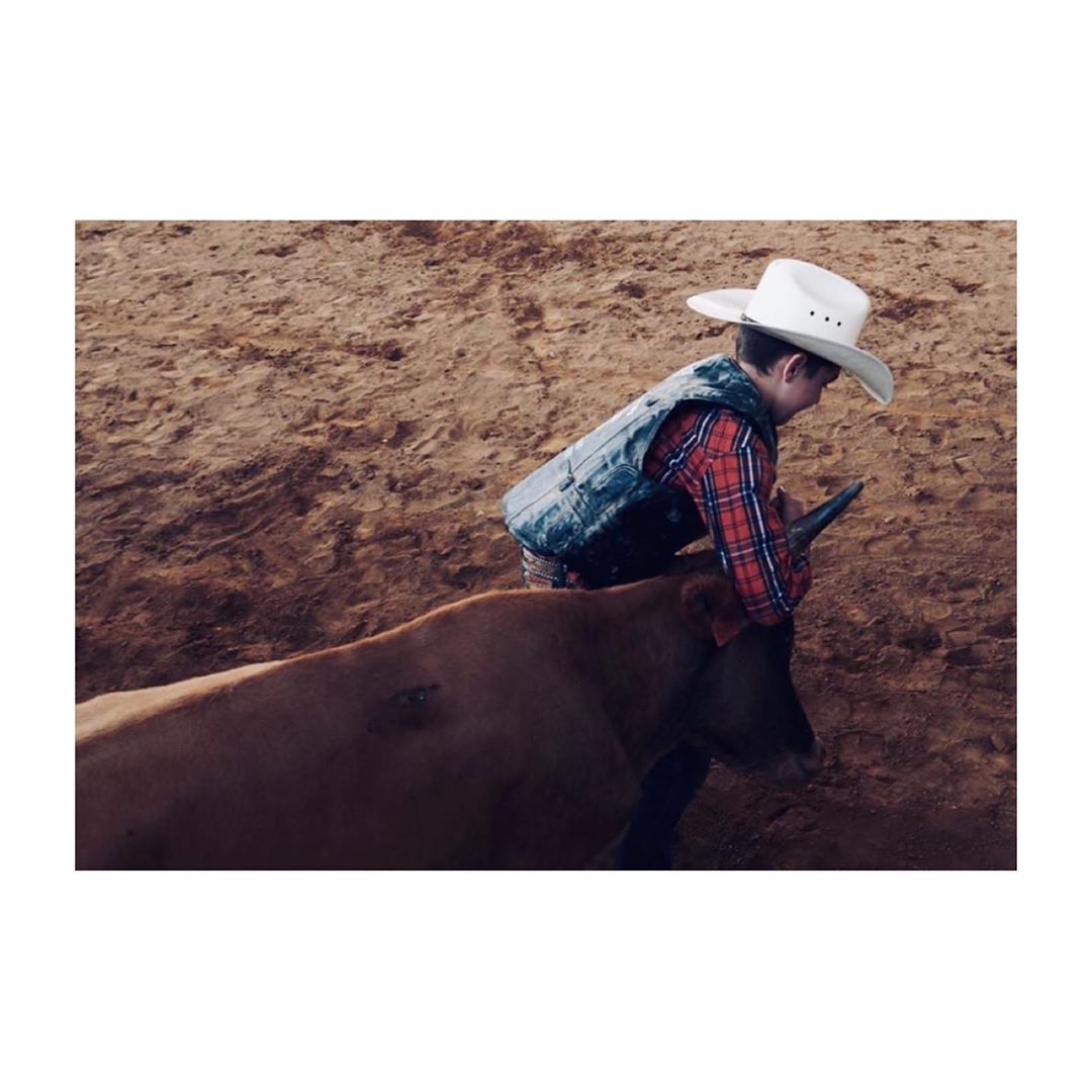 Young child rides a bull in a rodeo arena wearing a cowboy hat, vest, and plaid shirt in an eye-level medium shot.