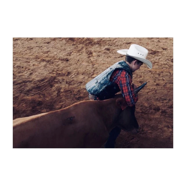 Young child rides a bull in a rodeo arena wearing a cowboy hat, vest, and plaid shirt in an eye-level medium shot.