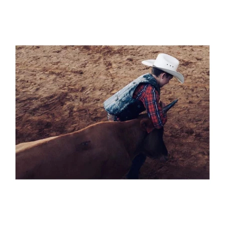 Young child rides a bull in a rodeo arena wearing a cowboy hat, vest, and plaid shirt in an eye-level medium shot.