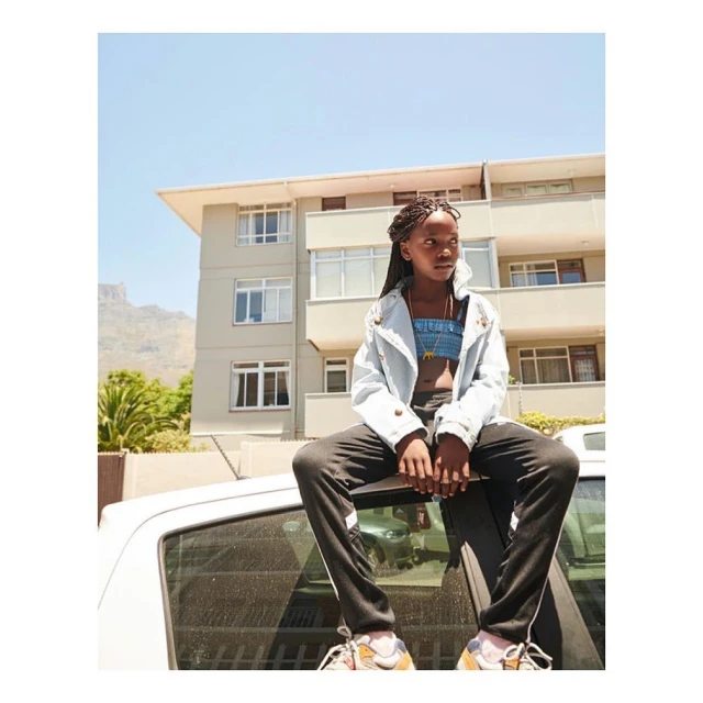 Young African-descent woman in braids sits on a car top in a city scene, wearing a light jacket, with apartment buildings behind.