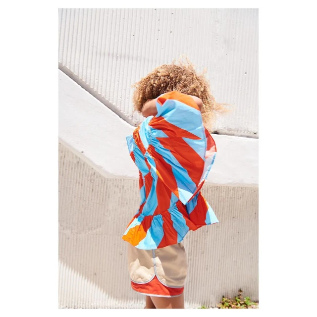 A child with curly hair wears a colorful, frilly top and shorts while standing against a textured, geometric white wall outdoors.