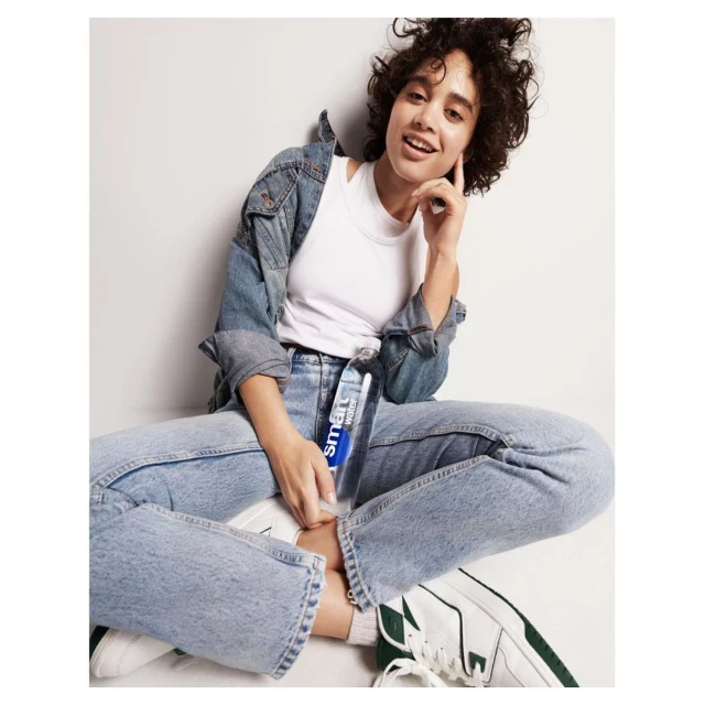 Young woman in denim jacket and jeans sitting cross-legged with bottled water in a bright studio, smiling and posing for a beauty, fashion ad.