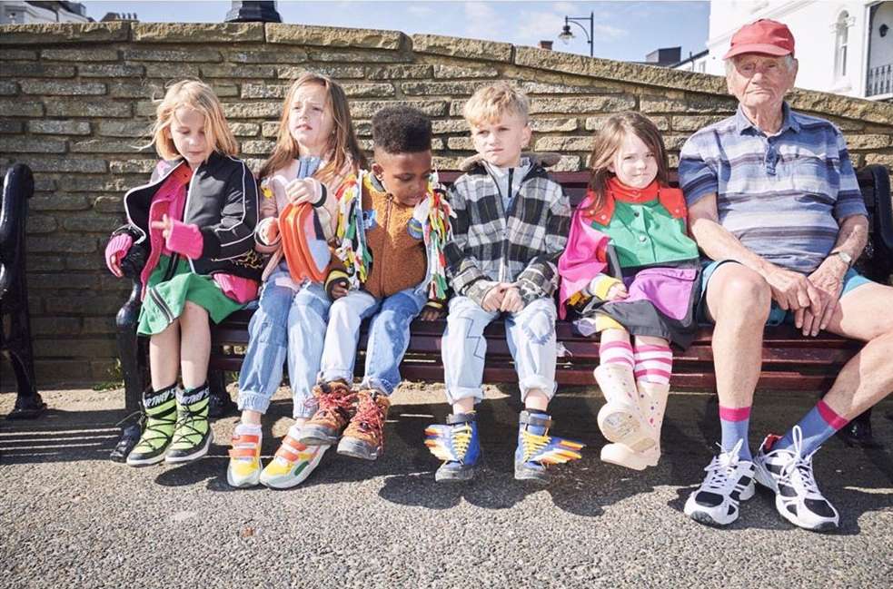 Group portrait featuring five young children and one elderly man seated on a park bench in casual wear with a stone wall background.