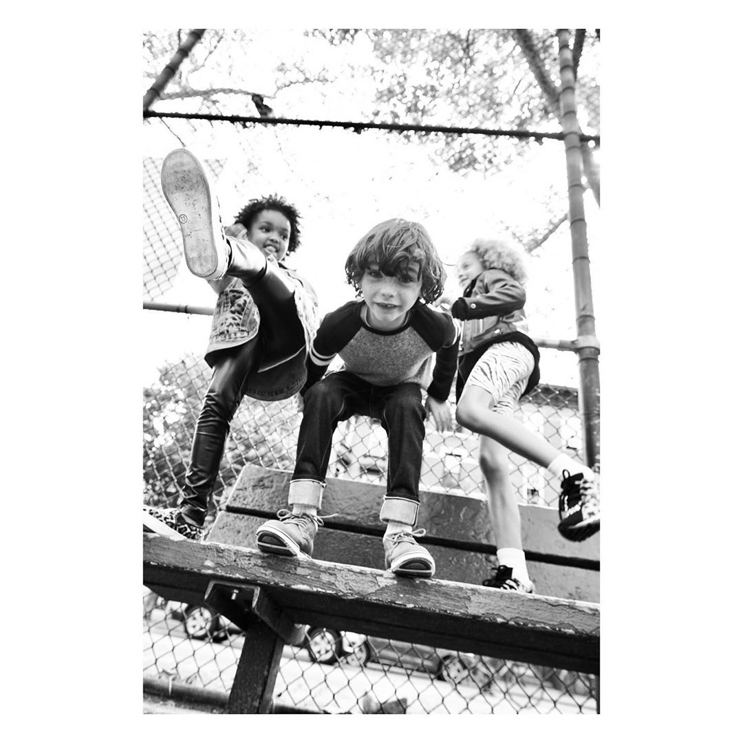 Three children, two girls and a boy, play on a wooden structure outdoors against a backdrop of greenery and a fence in black and white.
