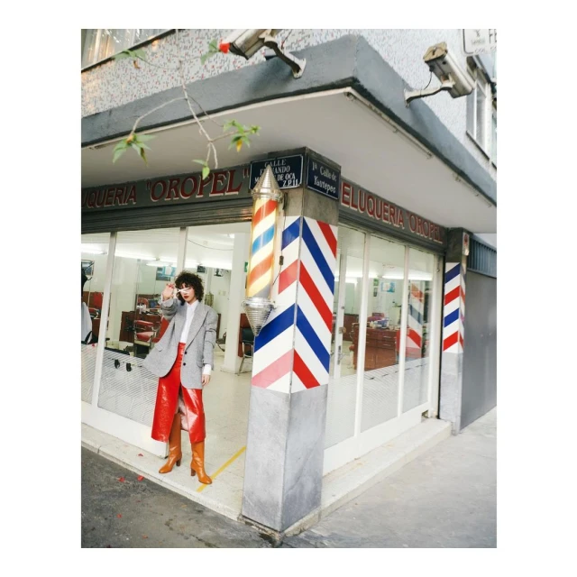 A young woman in a grey blazer and red pants poses in front of a barber shop with striped poles in an urban setting.