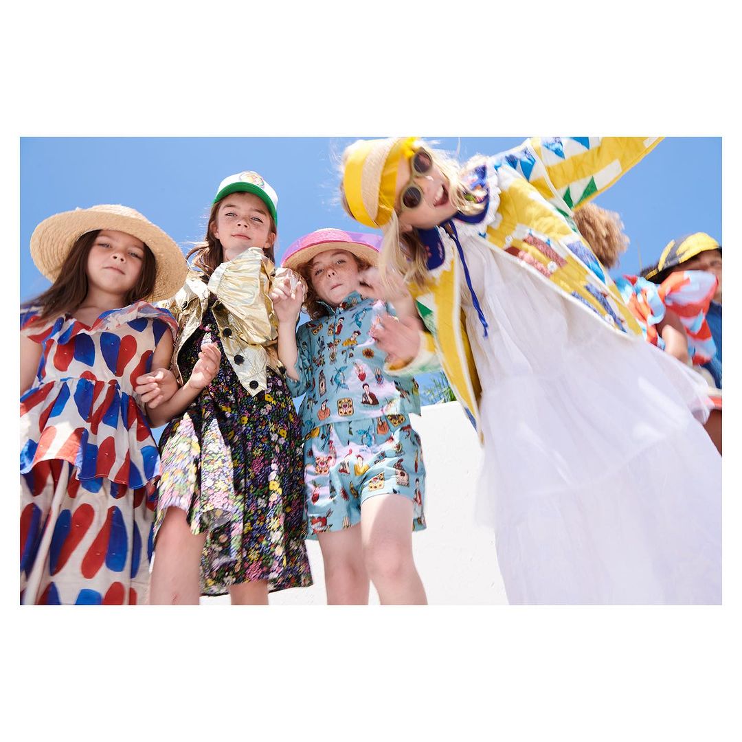 Four young girls in colorful outfits holding hands and looking at the camera under a blue sky in a low-angle outdoor shot.