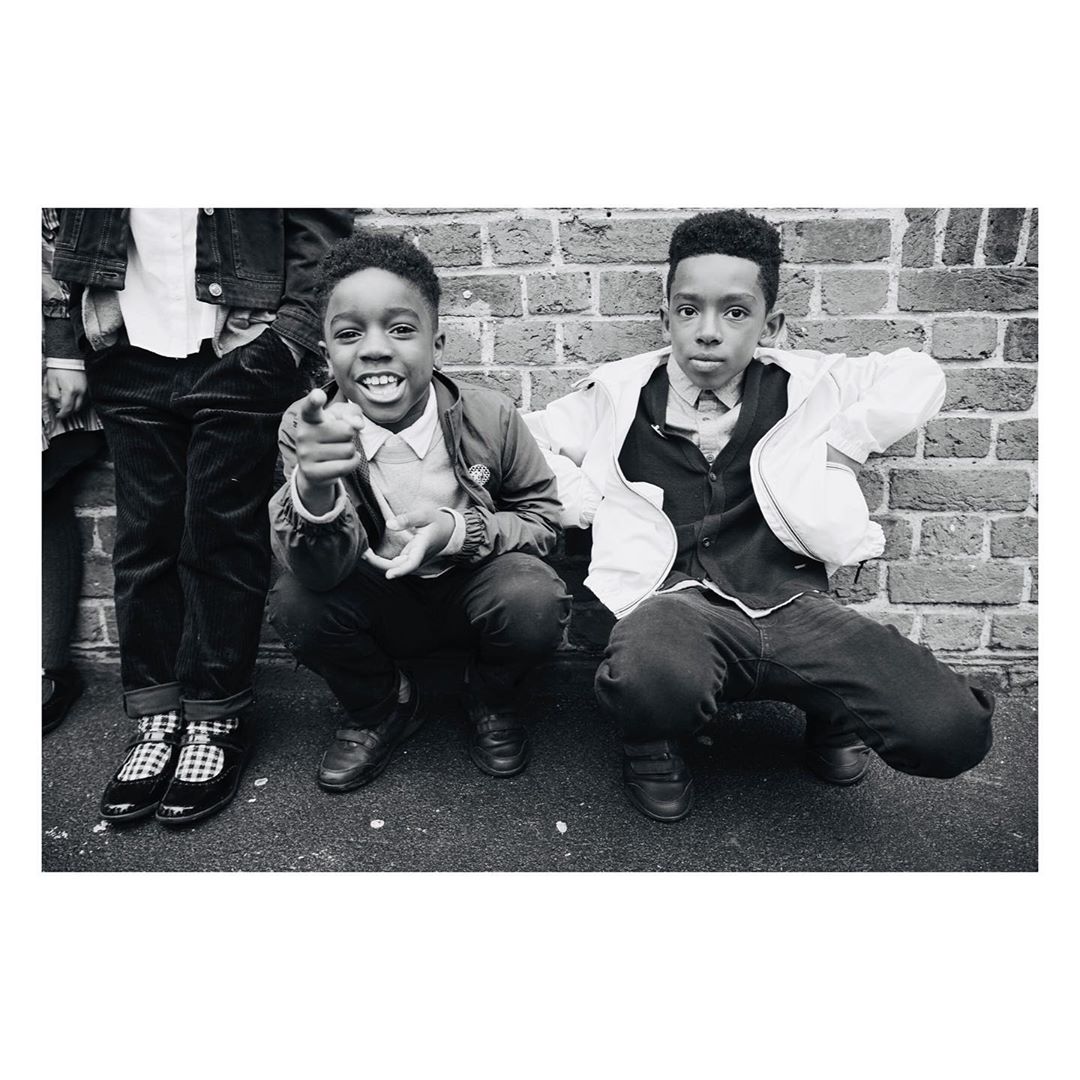 Black and white studio shot of two boys squatting in front of brick wall, smiling and pointing, wearing jackets and button-down shirts.