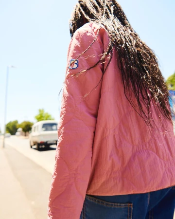 Rear view of a person with long braided dark hair wearing a pink quilted jacket walking down a sunny street.