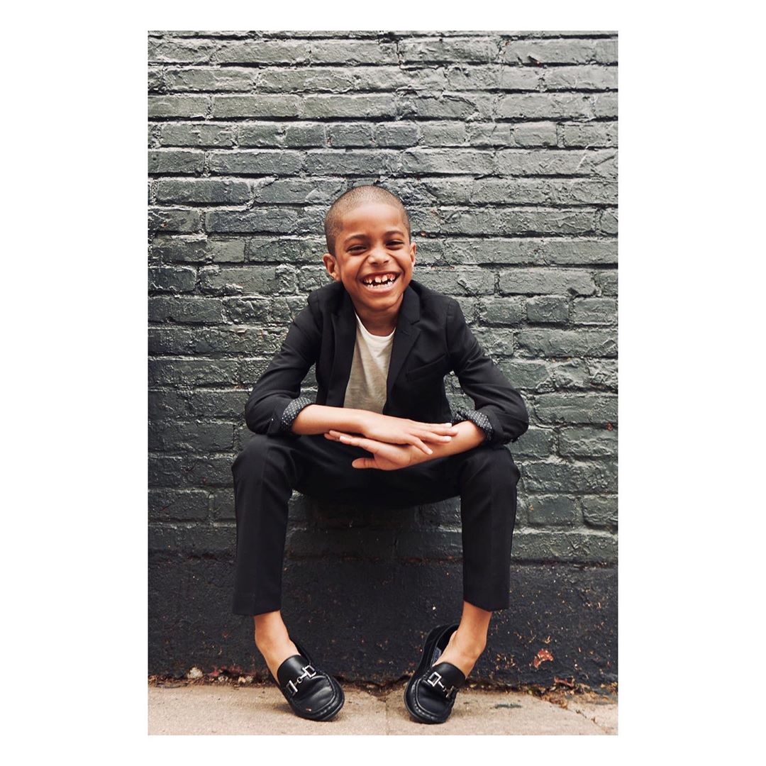 Young boy in a black suit and loafers sits against a gray brick wall, smiling broadly in an outdoor setting with natural light.