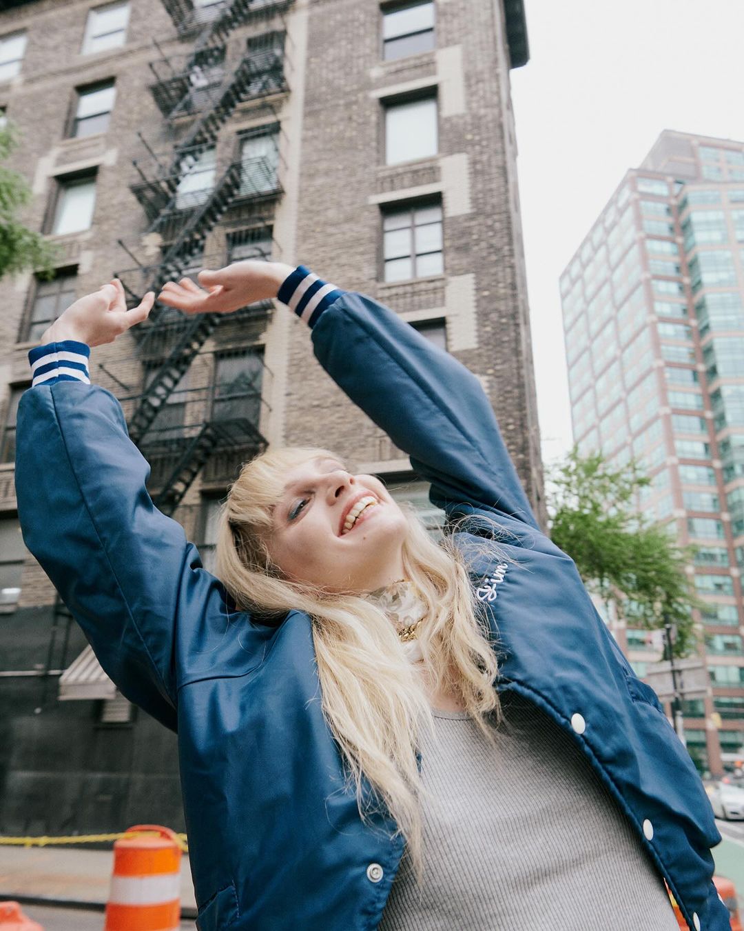 Smiling woman in blue jacket stands on city street, with brick building and modern skyscrapers in the background, reaching arms overhead.