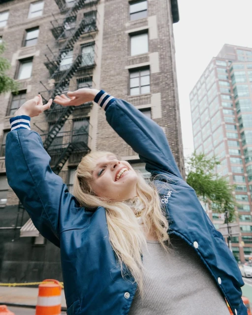 Smiling woman in blue jacket stands on city street, with brick building and modern skyscrapers in the background, reaching arms overhead.