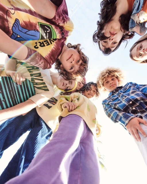 Group of children in trendy casual outfits looking down into the camera, forming a circle, low-angle view against clear sky. 