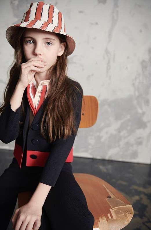 Child model poses seated in a striped hat, collared shirt, and dark suit against a neutral studio backdrop, showcasing fashion details.