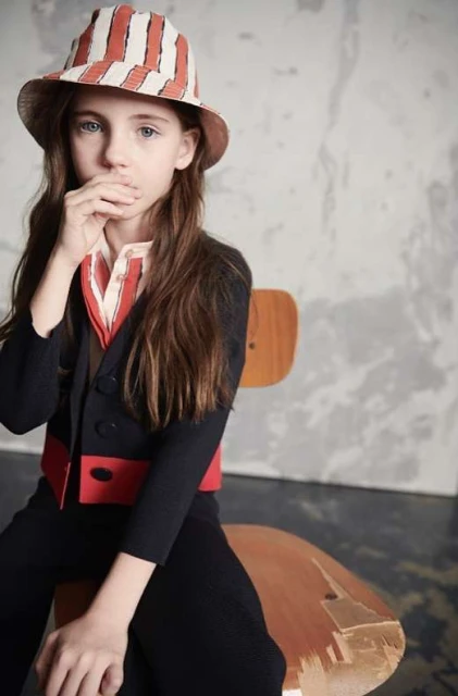 Child model poses seated in a striped hat, collared shirt, and dark suit against a neutral studio backdrop, showcasing fashion details.