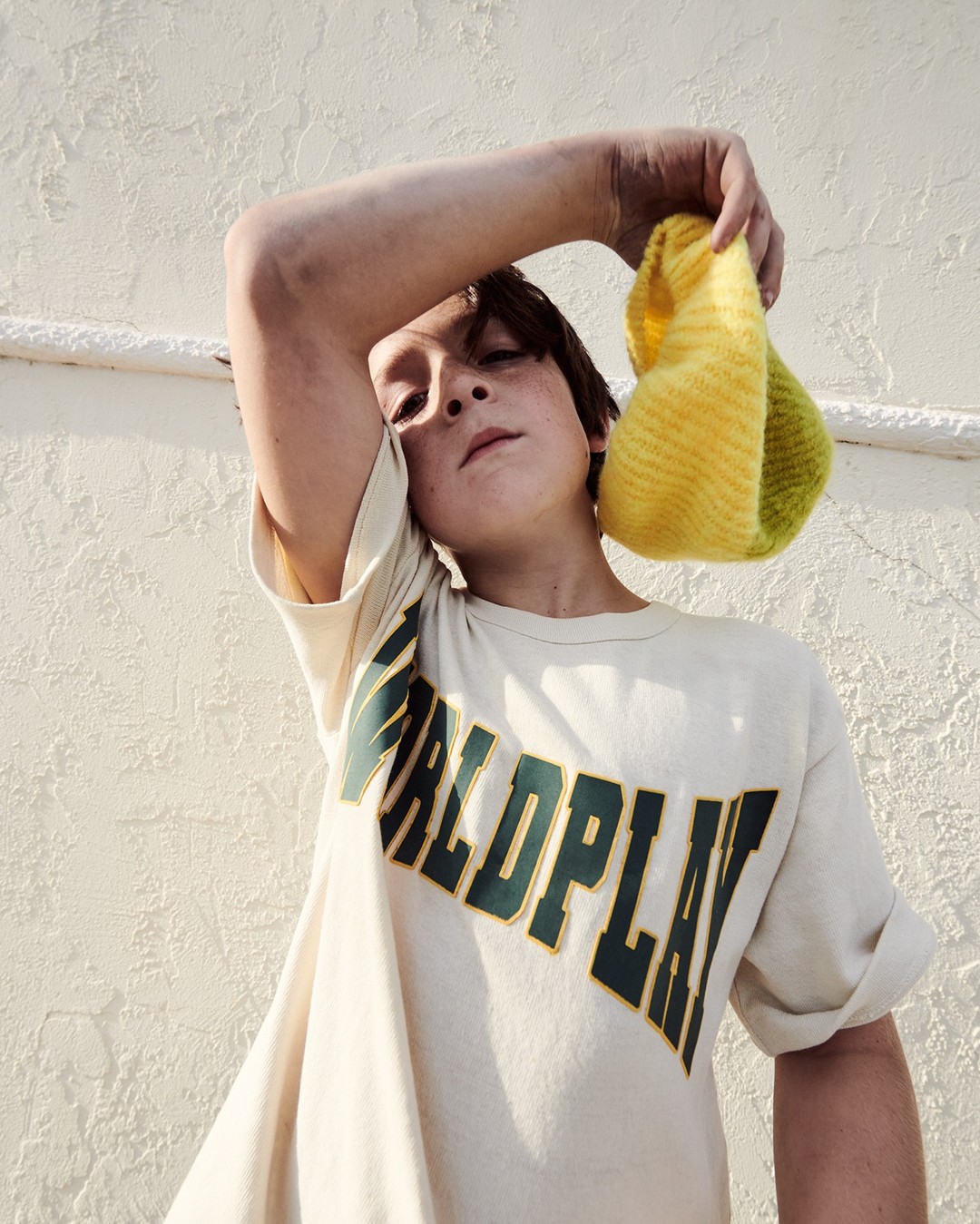 Teenager poses with a yellow and green knitted snood in a bright, naturally-lit indoor setting, wearing a 'Worldplay' graphic tee.