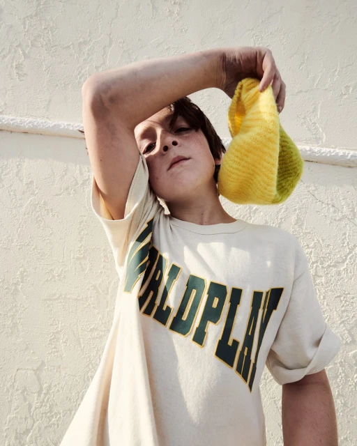 Teenager poses with a yellow and green knitted snood in a bright, naturally-lit indoor setting, wearing a 'Worldplay' graphic tee.