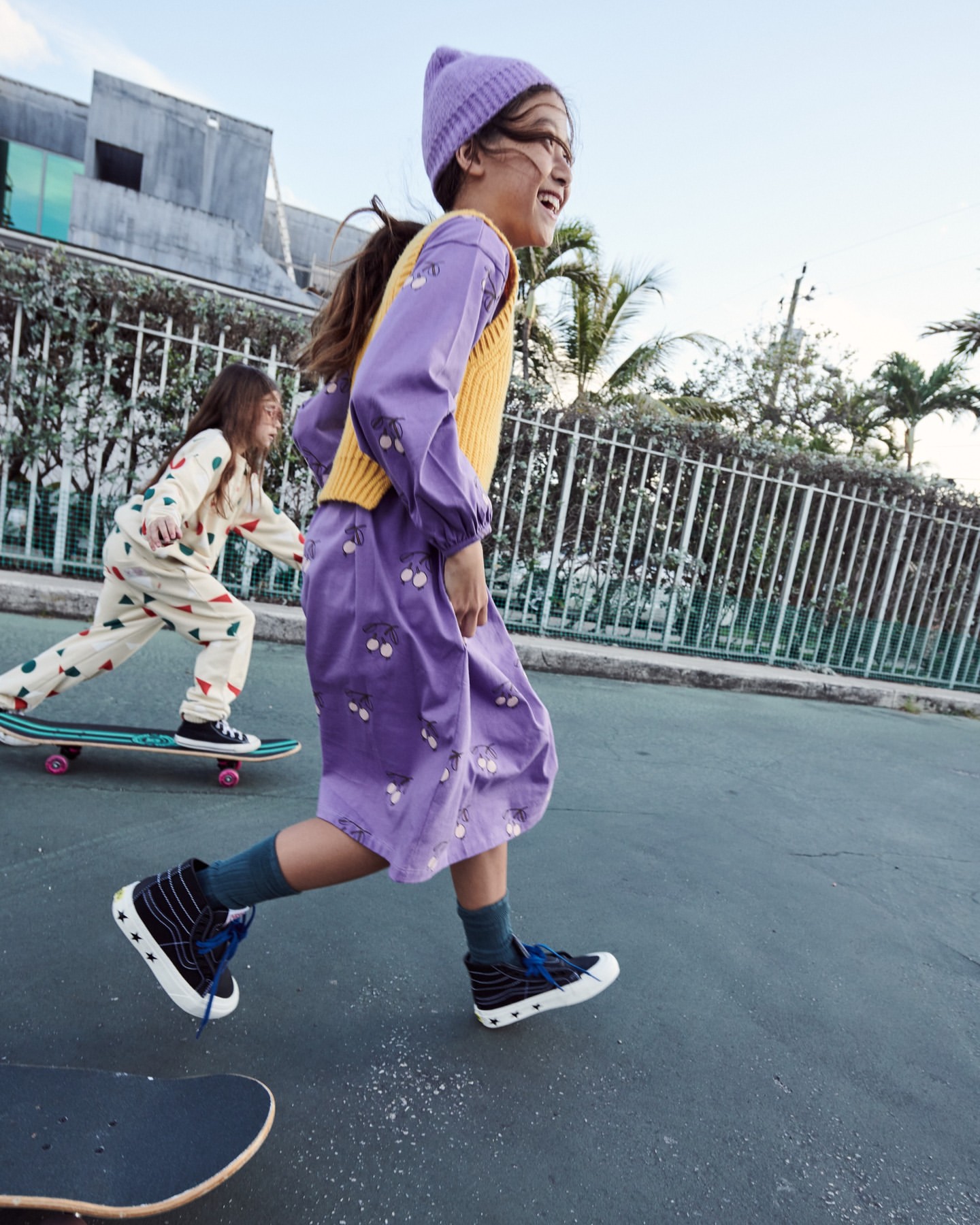Two young girls enjoying a sunny day outdoors, one running in a purple dress, the other skateboarding in a patterned outfit.