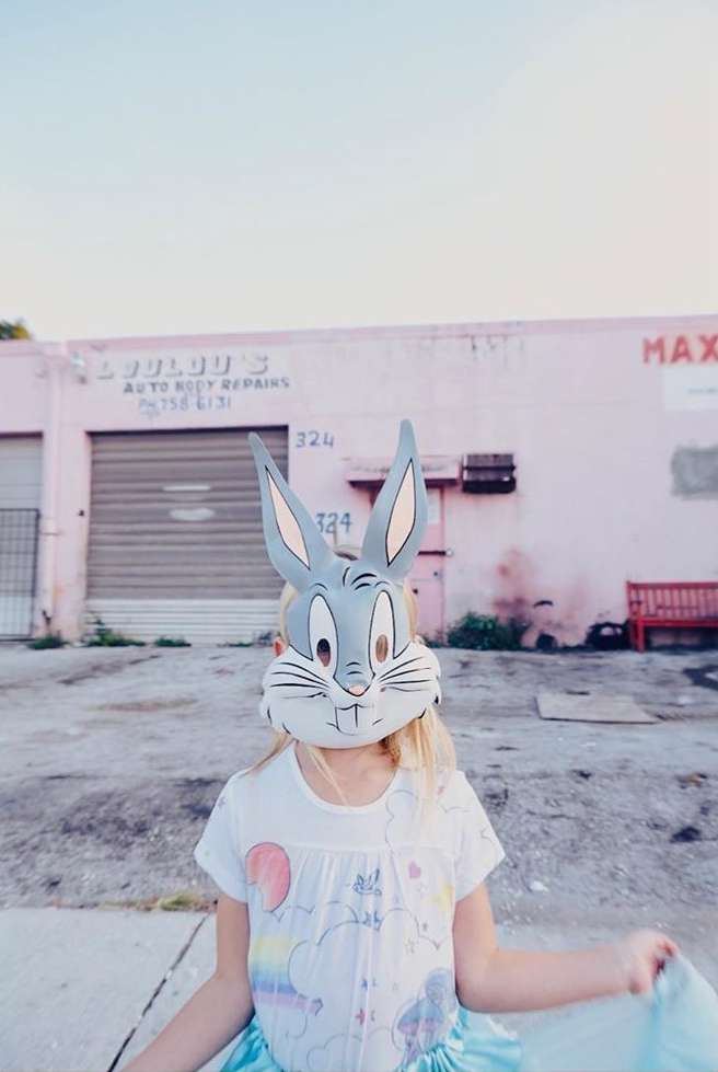 Young girl wearing Bugs Bunny mask in front of pink building with weathered exterior in a daylight outdoor shot.