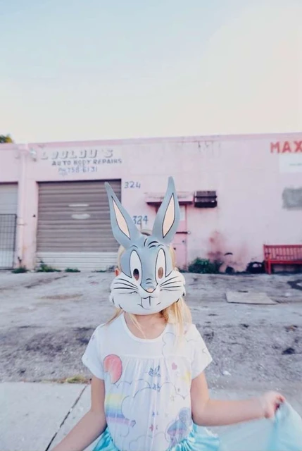 Young girl wearing Bugs Bunny mask in front of pink building with weathered exterior in a daylight outdoor shot.