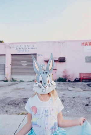 Young girl wearing Bugs Bunny mask in front of pink building with weathered exterior in a daylight outdoor shot.