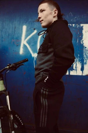 Young man in a black tracksuit standing beside a bicycle against a blue graffiti wall by Tim Marsella