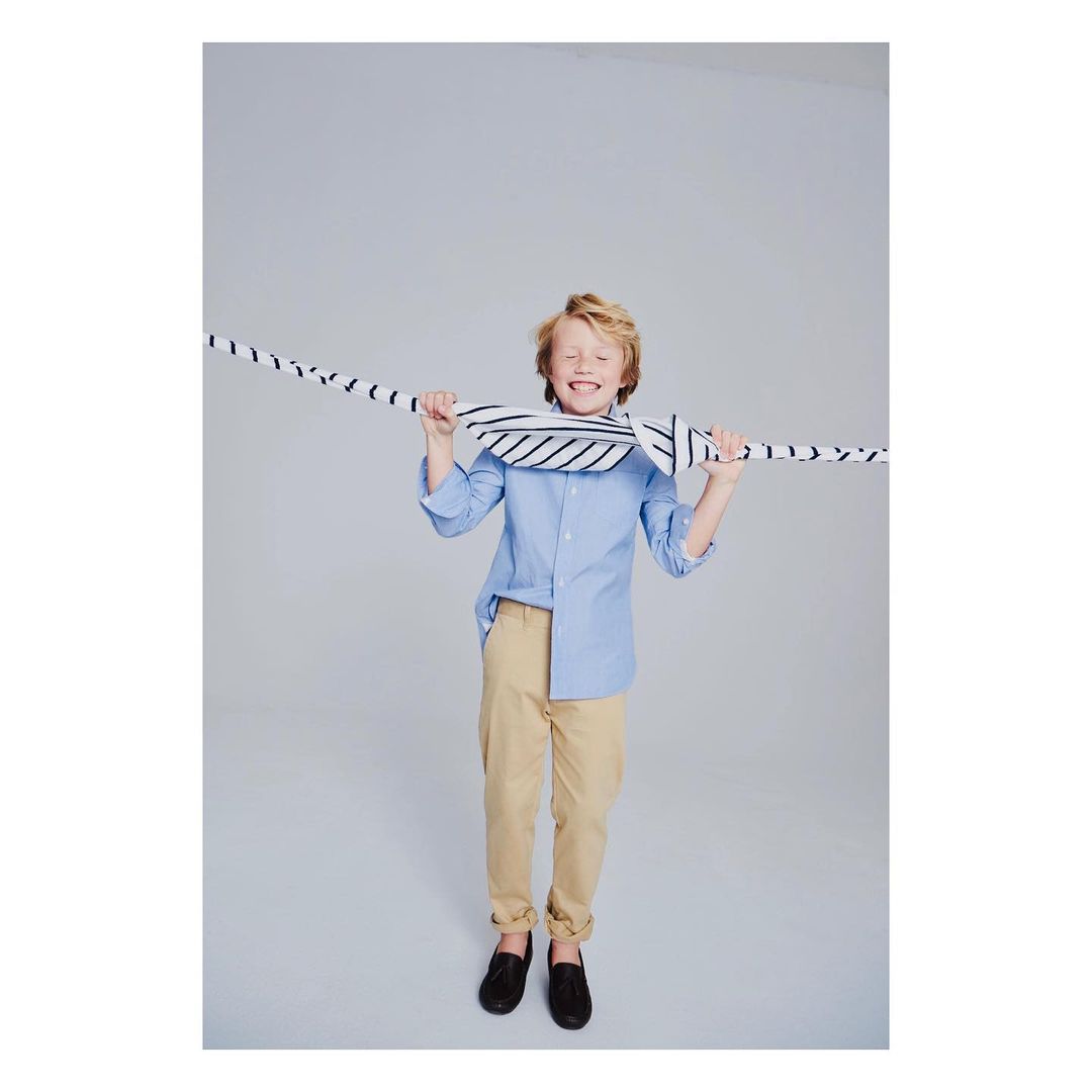 A smiling child in a light blue shirt and khaki pants stands against a gray backdrop, playfully holding a striped scarf over their head.