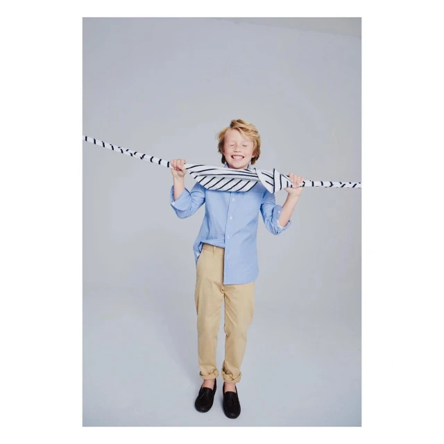 A smiling child in a light blue shirt and khaki pants stands against a gray backdrop, playfully holding a striped scarf over their head.