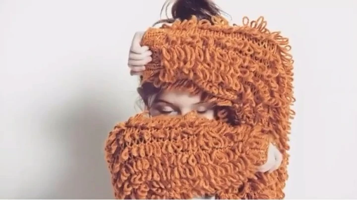 Woman wearing a heavy textured amber loop-knit sweater partially obscuring her face against a plain white background.