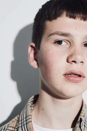 Close-up portrait of a young man wearing a plaid shirt and white t-shirt against a plain white studio background.