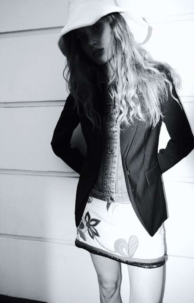 Model wearing a white bucket hat, dark blazer, and patterned skirt standing against a paneled wall.