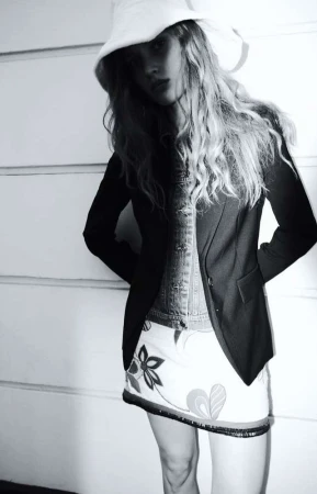 Model wearing a white bucket hat, dark blazer, and patterned skirt standing against a paneled wall.
