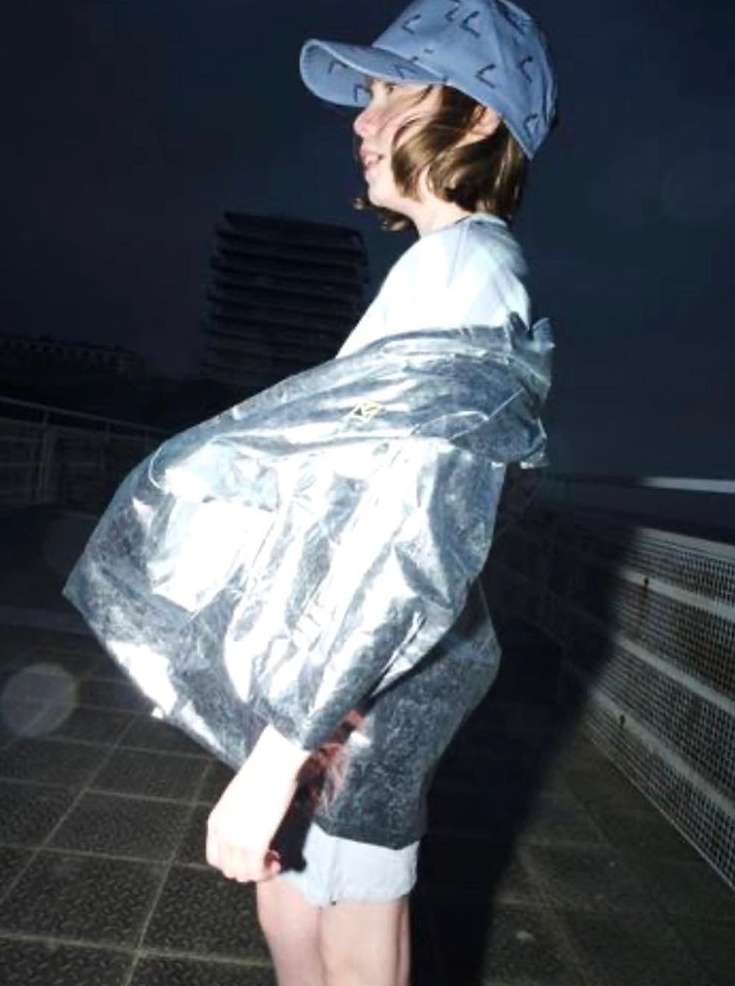 A young person wearing a light blue cap and a reflective metallic jacket stands outside outdoors at night.