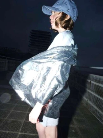 A young person wearing a light blue cap and a reflective metallic jacket stands outside outdoors at night.