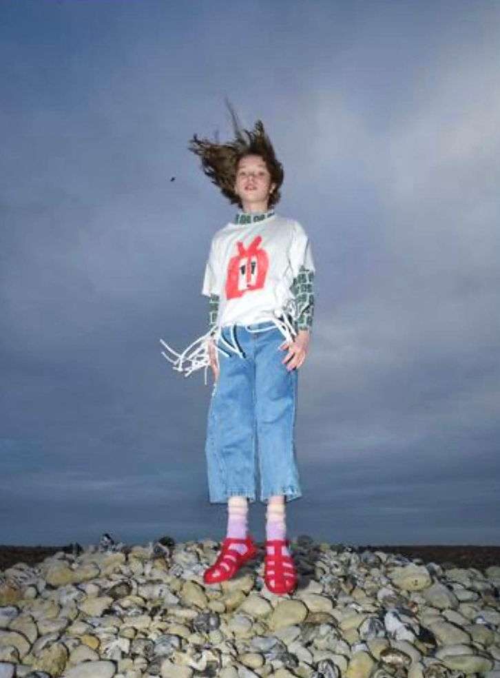 Full shot of a child in mid-air on a rocky beach, wearing a graphic tee, denim culottes, and red sandals, against a cloudy sky backdrop.