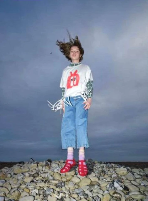 Full shot of a child in mid-air on a rocky beach, wearing a graphic tee, denim culottes, and red sandals, against a cloudy sky backdrop.