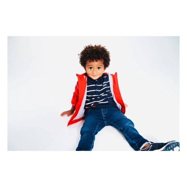 A young boy with curly hair sits on a white surface, wearing a red jacket, striped shirt, jeans, and sneakers, looking directly at the camera.