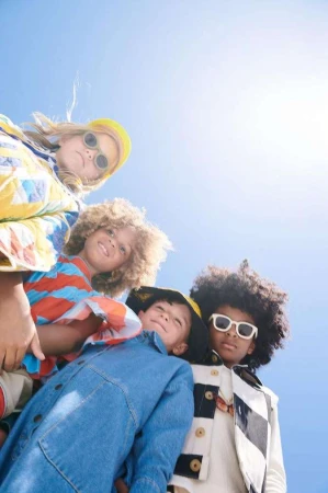 A group of four children, two girls and two boys, stand together under a clear sky, wearing bright and colorful clothing and sunglasses.