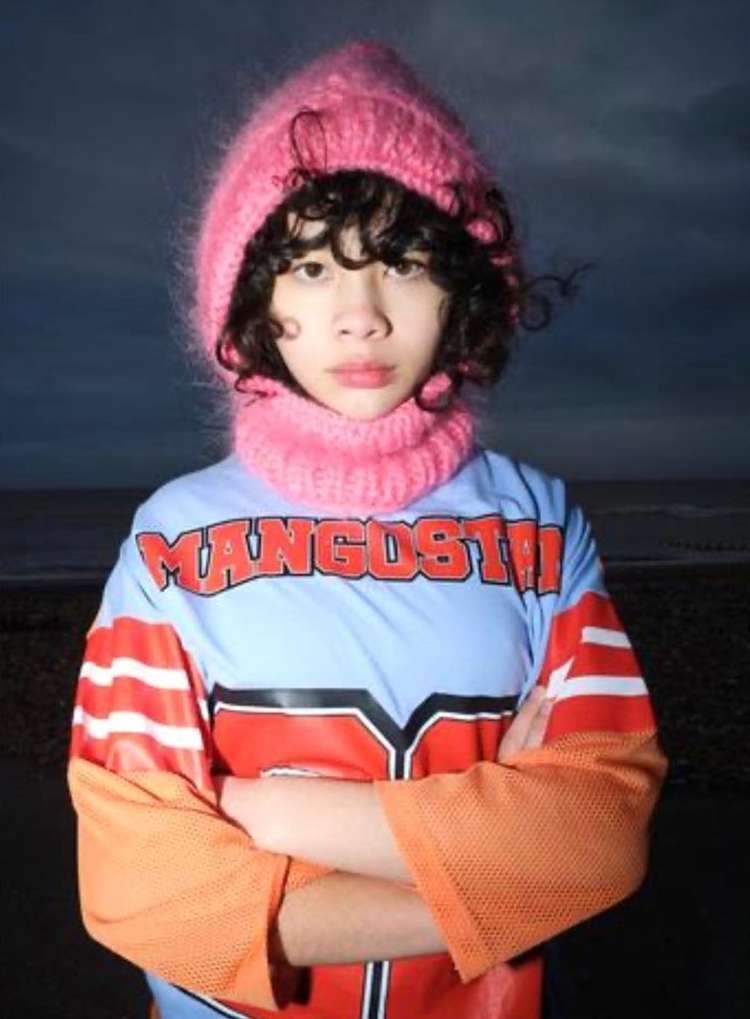 Teenager in a pink knit hat and neck warmer stands with arms crossed, wearing a color-blocked athletic jersey against an outdoor backdrop.