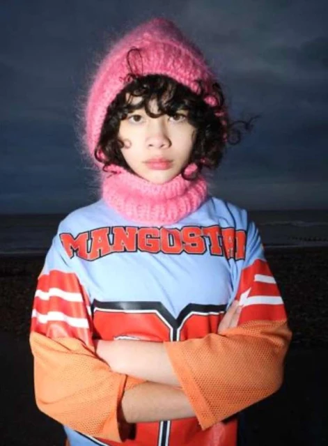 Teenager in a pink knit hat and neck warmer stands with arms crossed, wearing a color-blocked athletic jersey against an outdoor backdrop.