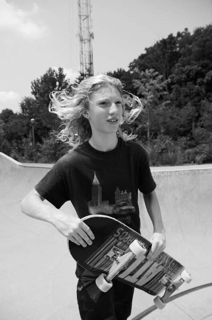 Teen skater holding a skateboard at an outdoor skatepark. Trees, sky, and a communications tower visible in the background.