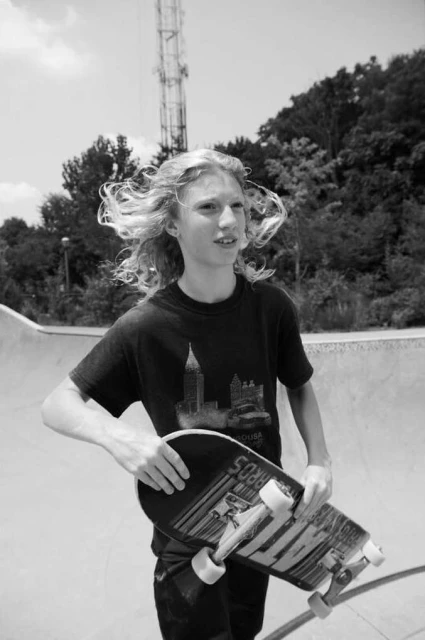 Teen skater holding a skateboard at an outdoor skatepark. Trees, sky, and a communications tower visible in the background.
