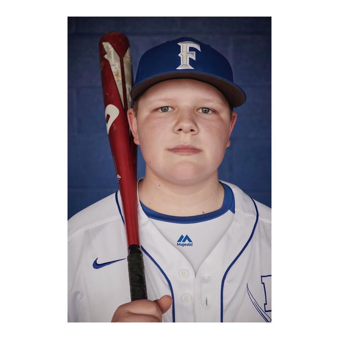 A young baseball player stands indoors wearing a white and blue uniform and a matching baseball cap, holding a baseball bat in a portrait shot.