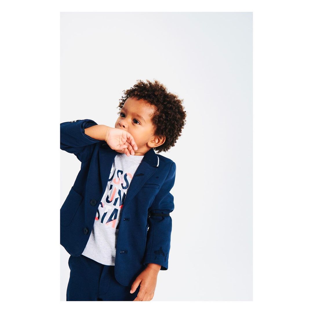 Young child in a navy blazer and matching pants stands in a photo studio with his fingers near his mouth looking up.