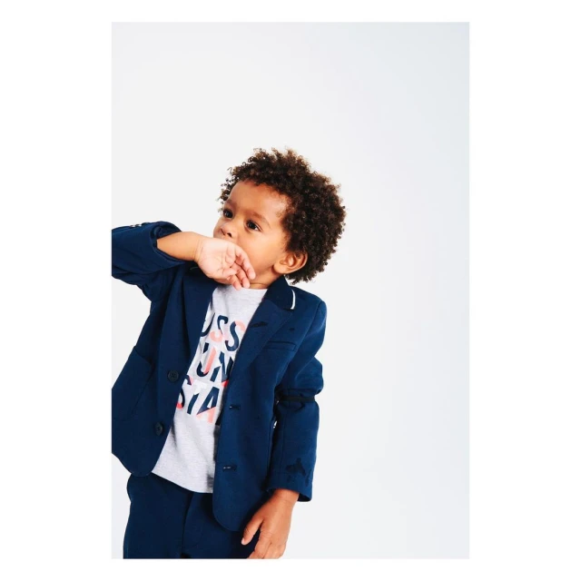 Young child in a navy blazer and matching pants stands in a photo studio with his fingers near his mouth looking up.