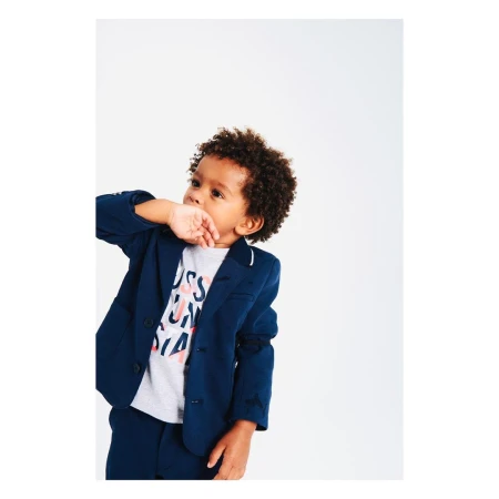 Young child in a navy blazer and matching pants stands in a photo studio with his fingers near his mouth looking up.