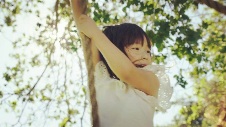 Child in a white dress hugs a tree in a sun-drenched forest, smiling and enjoying the natural setting with lush green leaves.