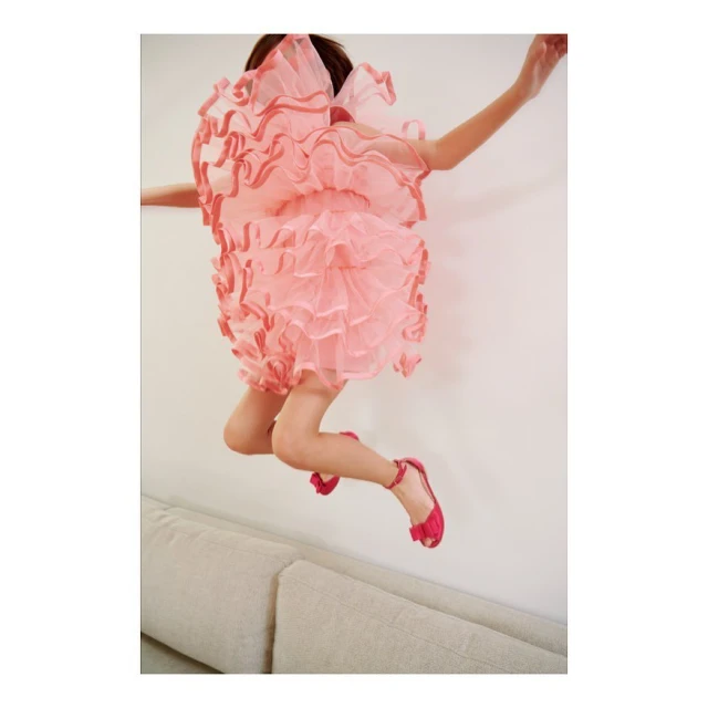 A child jumps on a couch in a pink, frilly dress and matching pink shoes against a neutral wall, capturing a moment of playful energy and joy.