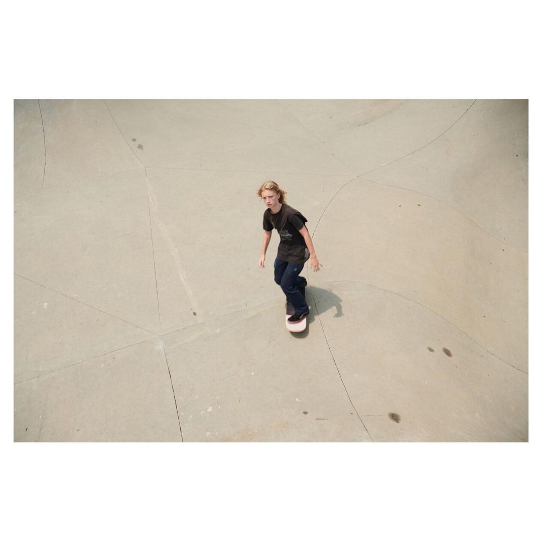 Young adult with long blond hair rides a skateboard in an outdoor concrete skatepark, shot from a high angle, capturing the sport's action.